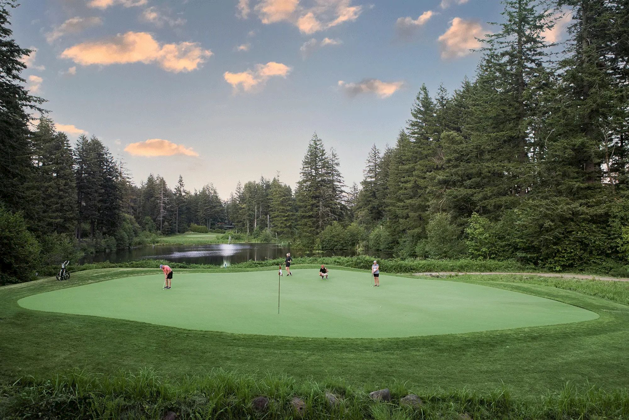 A golf course surrounded by trees with people playing on the green under a partly cloudy sky at sunset.