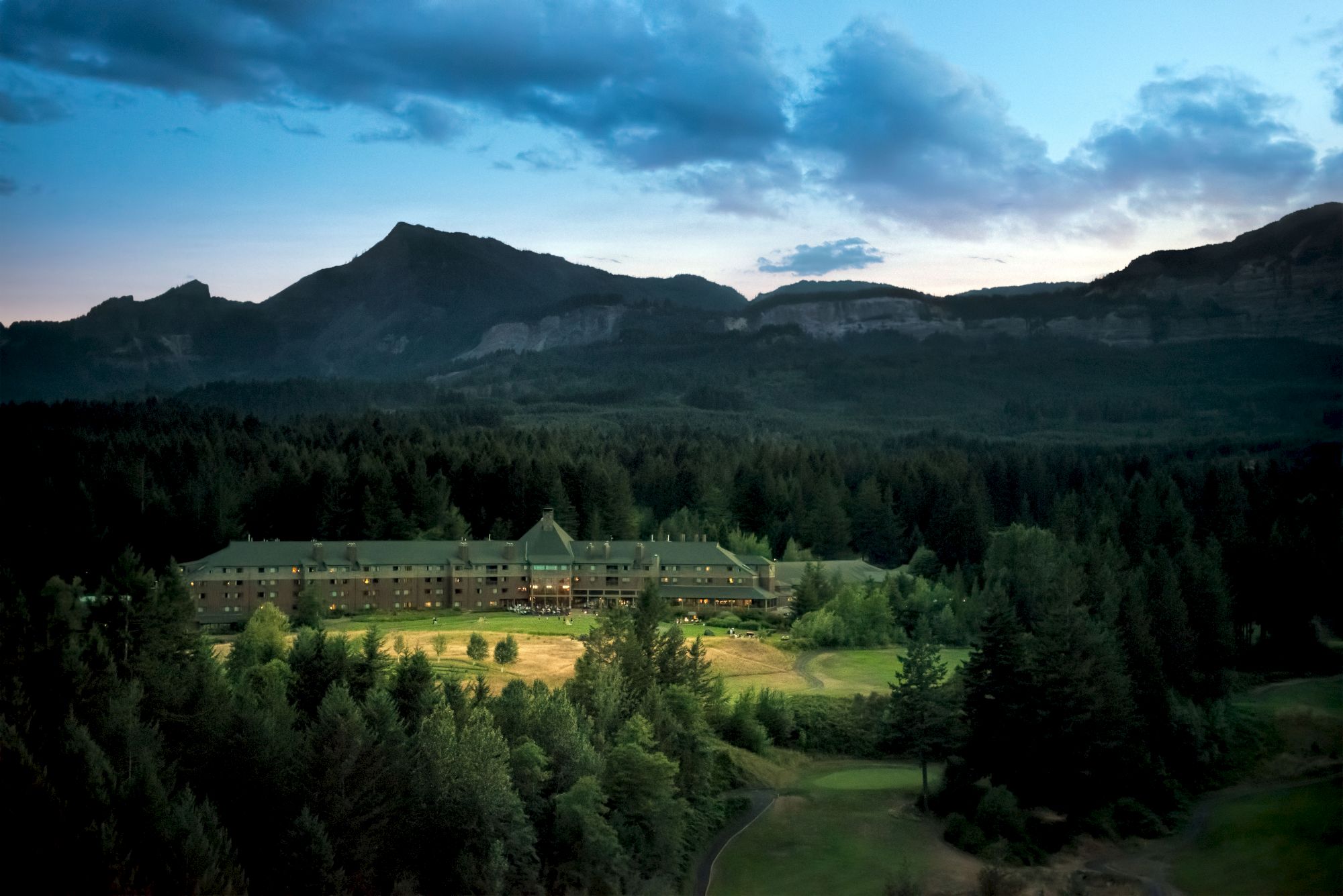 A building nestled among trees with mountains in the background at dusk under a partially cloudy sky.