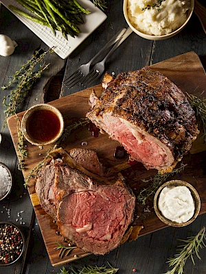 A cooked prime rib on a wooden board with gravy, horseradish, mashed potatoes, and roasted asparagus on the side, garnished with herbs.