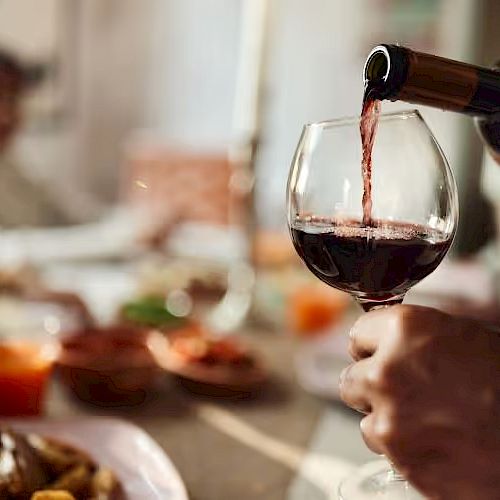 A person is pouring red wine into a glass at a dining table, with blurred figures and food in the background, creating a cozy atmosphere.