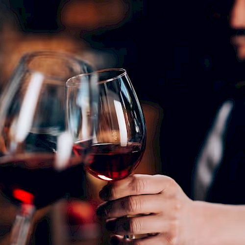 Several glasses of red wine are being clinked together in a toast, held by a person in a blurred background setting.