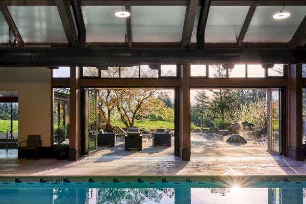 The image shows an indoor pool area with open glass doors leading to an outdoor seating area and garden, bathed in sunlight.