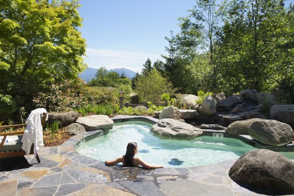 A person relaxes in a small stone-bordered pool surrounded by nature, trees, and distant mountains on a sunny day.