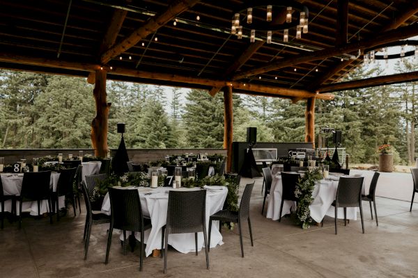 An outdoor event setup with round tables, white tablecloths, and black chairs under a wooden pavilion surrounded by trees in the background.