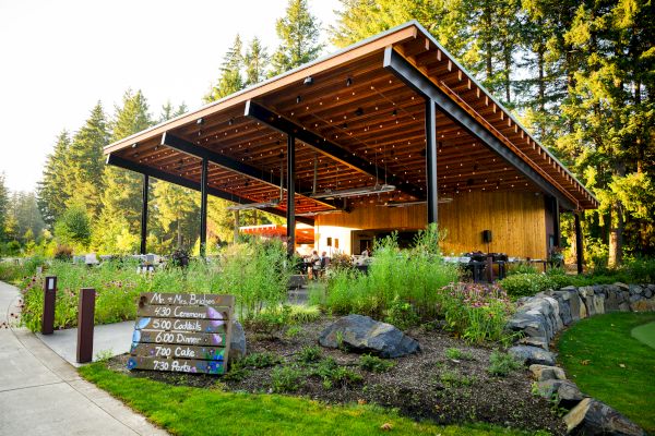 An outdoor venue with a wooden roof and string lights, surrounded by greenery. A schedule on a sign lists events like dinner and live music.