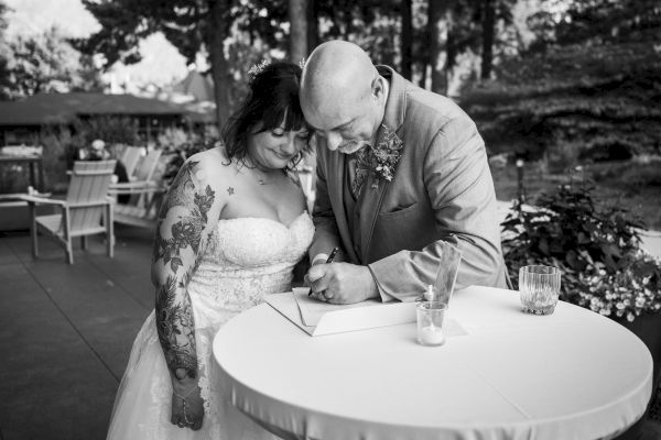 A couple signs a document on a round table at an outdoor event, possibly a wedding; they appear joyful and close.
