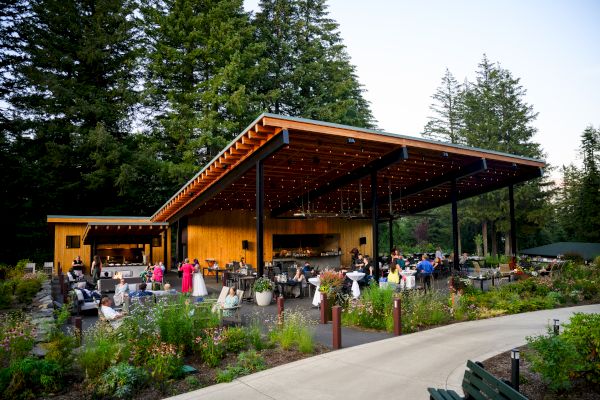 An outdoor restaurant with a wooden roof, surrounded by greenery and people dining and socializing.