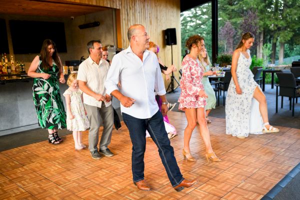 A group of people dancing indoors on a wooden floor, dressed in summer outfits, with a bar and outdoor view in the background.