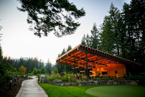 A pavilion with warm lighting in a wooded area, surrounded by trees and greenery, with a path leading towards it at dusk.