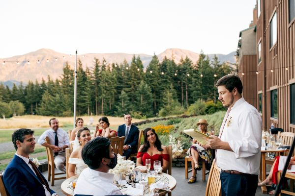 A group of people sit outside at wooden tables, listening to a man giving a speech. Trees and mountains are in the background.
