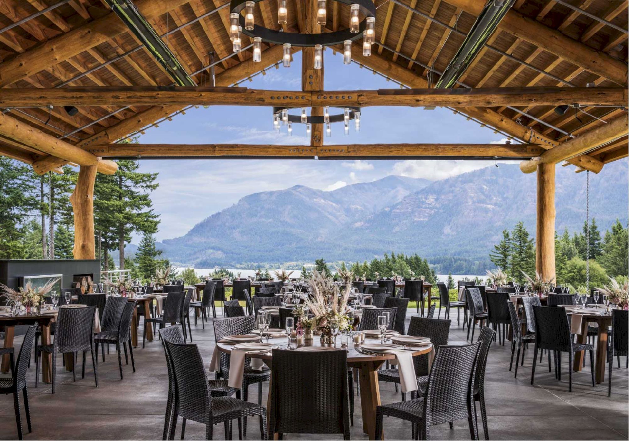 An elegant outdoor restaurant setup with wooden beams and mountain views. Round tables are set with tableware and floral centerpieces.