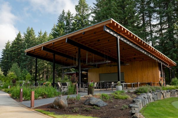 A modern open-air pavilion with a wooden roof is set in a lush, forested area with outdoor seating and landscaped surroundings.