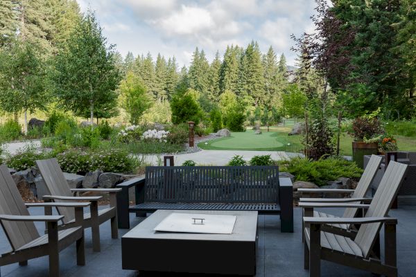A serene patio setup with wooden chairs and a fire pit, overlooking a lush garden and dense forest in the background.
