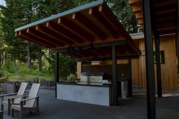 Outdoor kitchen with a wooden canopy, a grill, a countertop, and chairs, surrounded by greenery and a wooden building.