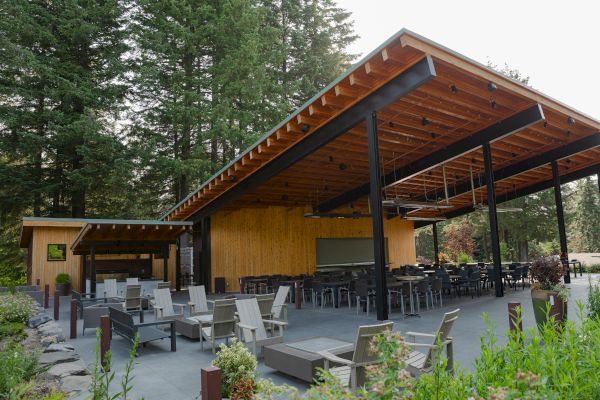 An outdoor seating area with wooden and metal structures, surrounded by lush greenery and trees, featuring chairs and tables.