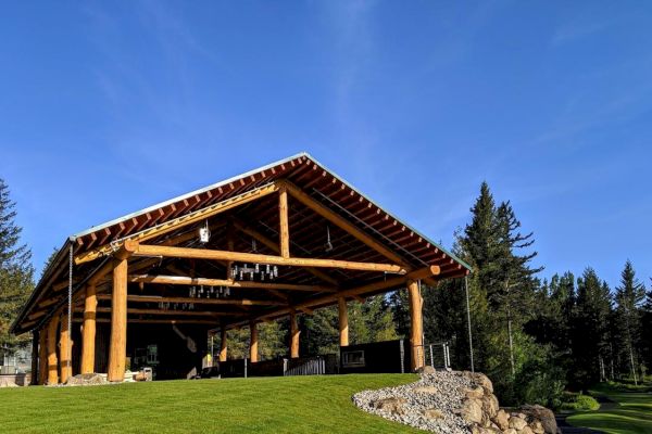 A large wooden pavilion with a sloped roof and beams stands on a grassy hill, surrounded by trees under a clear blue sky.