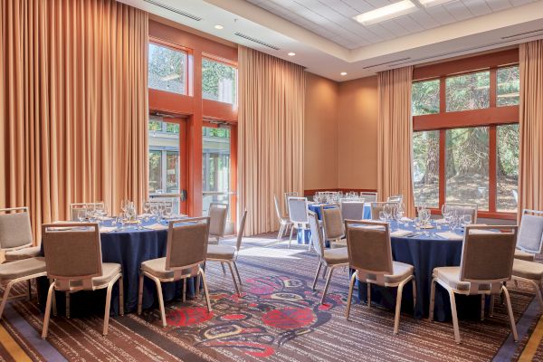 The image shows a banquet room with round tables set with blue tablecloths and chairs, surrounded by large windows and beige curtains.