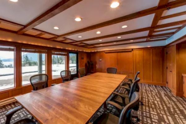 A conference room with wooden walls and ceiling, a large table, and chairs. Windows show an outdoor snowy landscape under a clear sky.