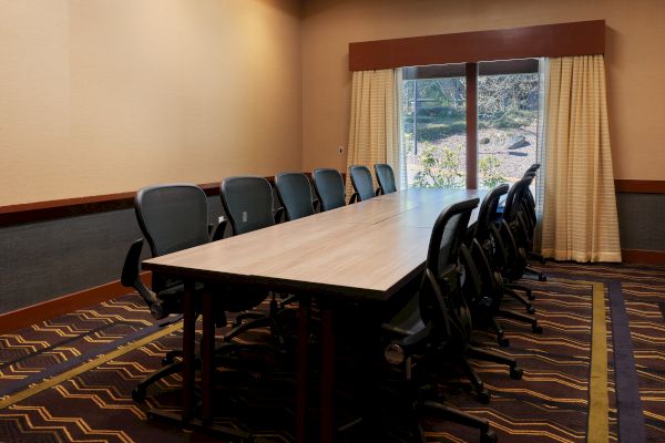 A conference room with a long table, black chairs, patterned carpet, and curtains over a large window in a neutral-toned room.