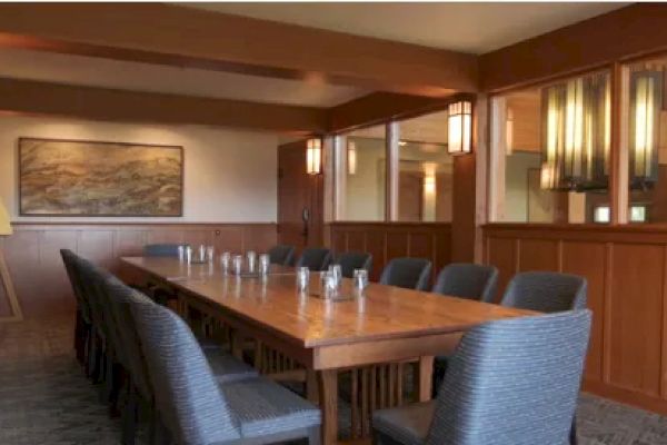 A long wooden conference table with several upholstered chairs is in a room with warm lighting and framed artwork on the wall.