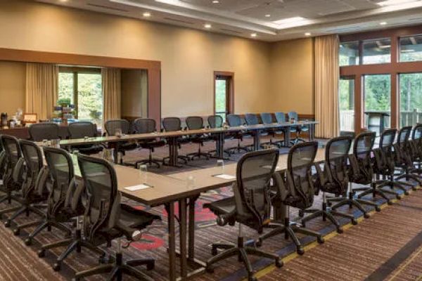 A large conference room with U-shaped tables, office chairs, and a beverage station, surrounded by windows with natural light.