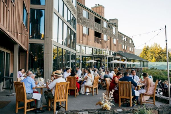 People are dining outdoors at a restaurant with a modern wooden exterior, enjoying a pleasant setting under string lights.