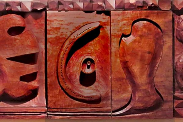 The image shows a wooden sculpture featuring abstract shapes and carved human faces, arranged in a series of panels.