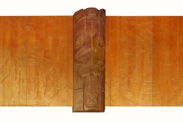 The image shows a wooden artwork with intricate carvings, featuring a central totem-like structure surrounded by engraved panels.