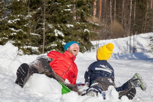 Two people in colorful clothing are playing and laughing in the snow against a backdrop of trees.