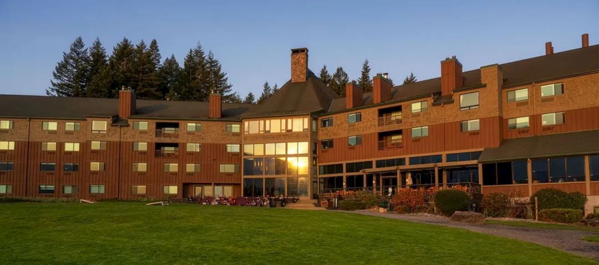 A large building with wooden and brick elements, surrounded by green grass and trees, under a clear blue sky at sunset.