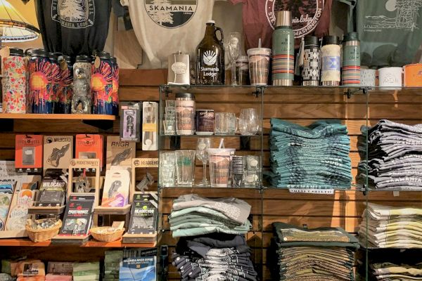 This image shows a retail display with T-shirts, water bottles, postcards, books, candles, and stacks of towels or clothes on shelves.