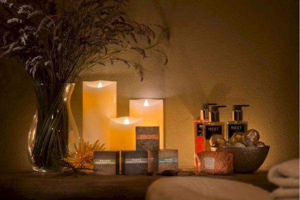 The image shows a cozy spa setting with lit candles, lotions, aromatherapy soaps, a vase of dried flowers, and folded towels on a table.