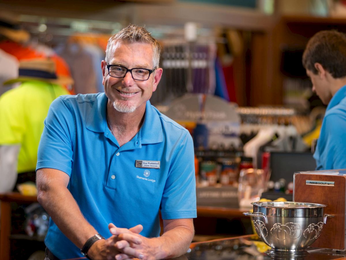 A smiling man in a blue polo shirt is standing in a shop, with another person working in the background, surrounded by merchandise displays.