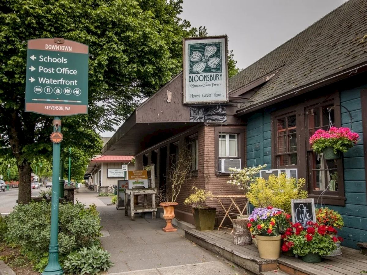 A quaint street with a flower shop adorned with plants and colorful blooms, and a signpost pointing to schools, the post office, and waterfront.