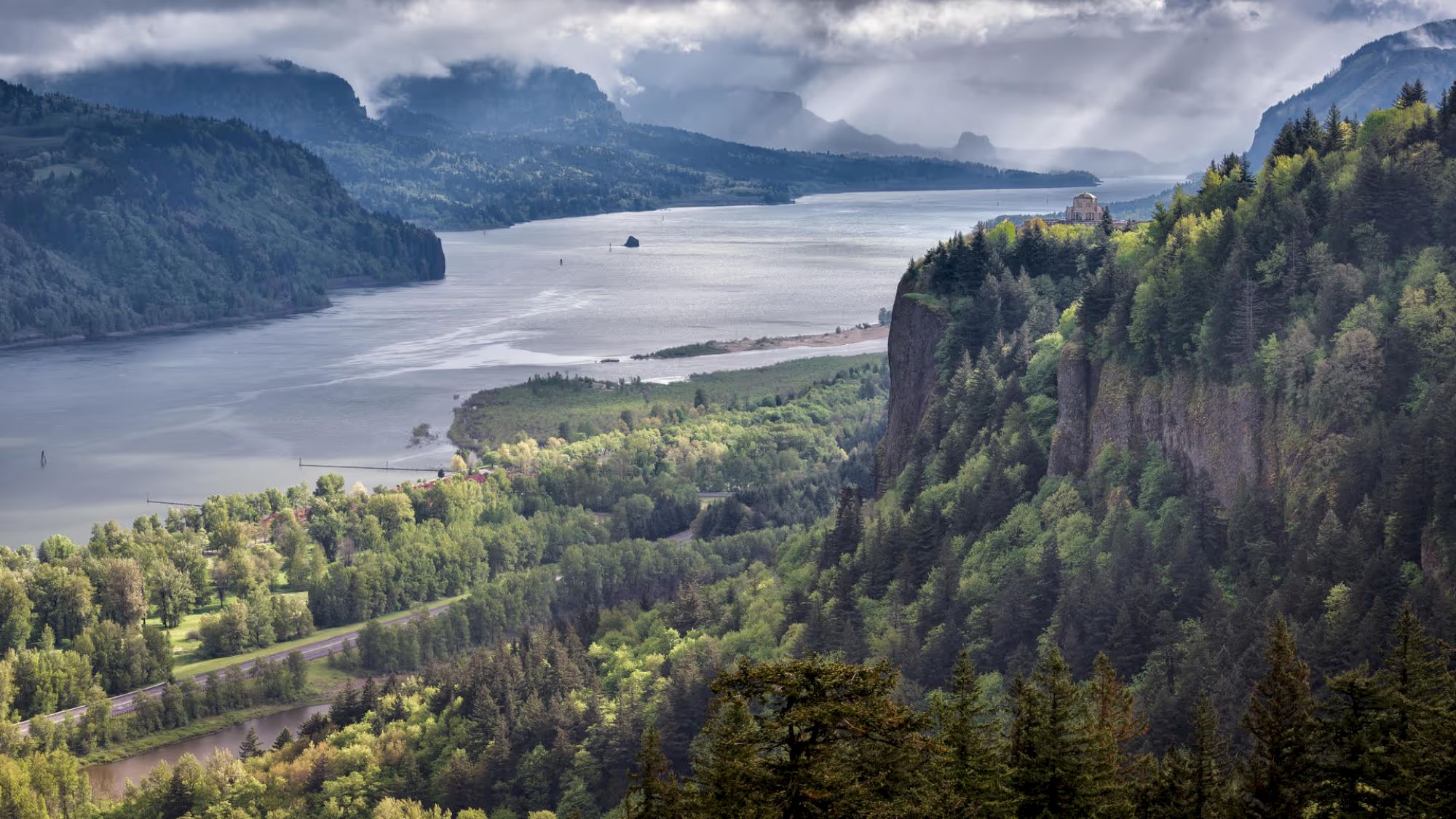 Scenic view of a river winding through a lush, forested valley with cliffs and mountains in the background under a cloudy sky.