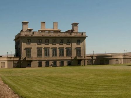 A large, historic building with multiple chimneys and expansive windows, surrounded by a well-maintained grass lawn under a clear sky.