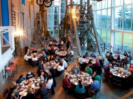 People are dining in a large, industrial-style space with high ceilings and unique decor, featuring eclectic elements and natural lighting.