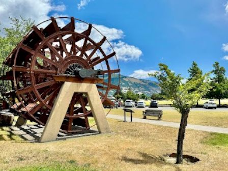 An old wooden water wheel sculpture stands in a park, with trees, cars, and mountains in the background under a clear blue sky.