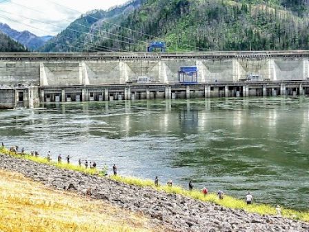 A dam spans a river, with people walking along its grassy bank. A mountain backdrop completes the scenic view.