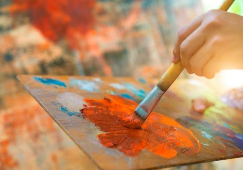 A hand holds a brush painting with orange paint on a palette, with a colorful background in soft focus.