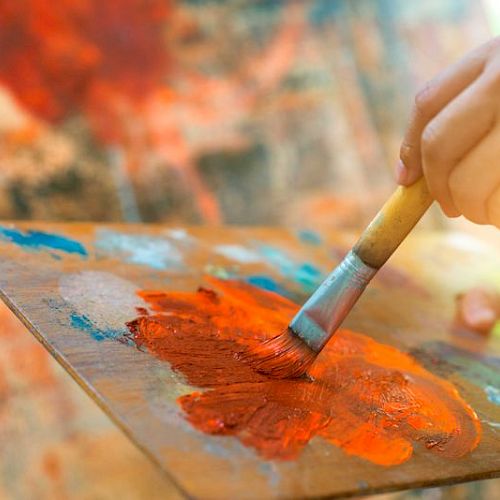 A hand holds a brush painting with orange paint on a palette, with a colorful background in soft focus.