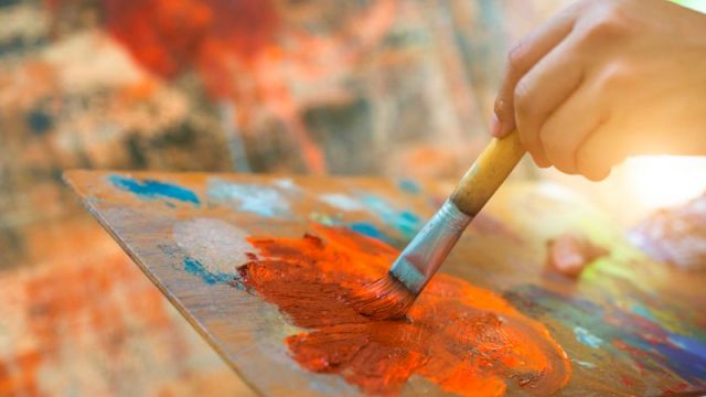 A hand holds a brush painting with orange paint on a palette, with a colorful background in soft focus.