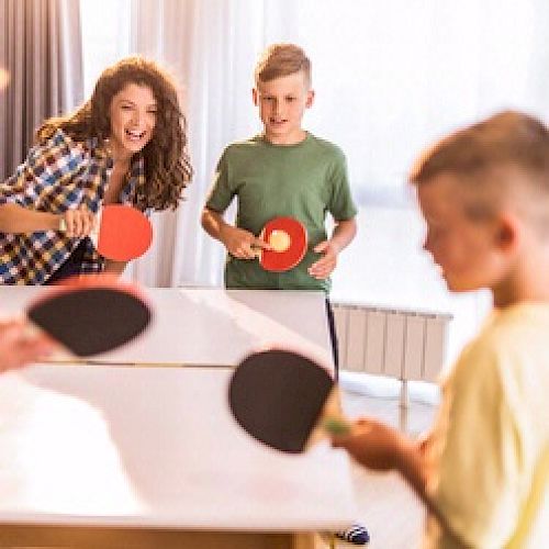 People are playing a doubles ping pong game indoors, appearing engaged and having fun with bright lighting in the background.