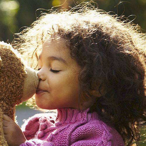 A child in a purple sweater is affectionately kissing a brown teddy bear in a sunlit outdoor setting.