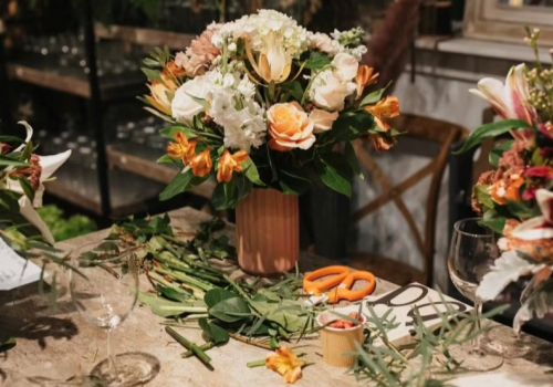A flower arrangement with lilies and roses is on a table, with scissors and wine glasses nearby, set in what seems like a cozy space.
