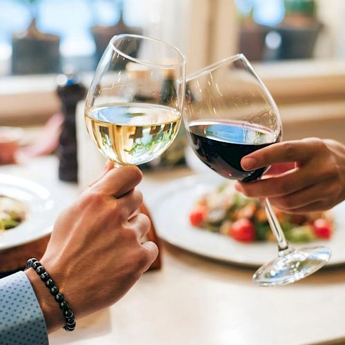 Two people clinking glasses of white and red wine over a dinner table with plates of food in the background, in a cozy setting.