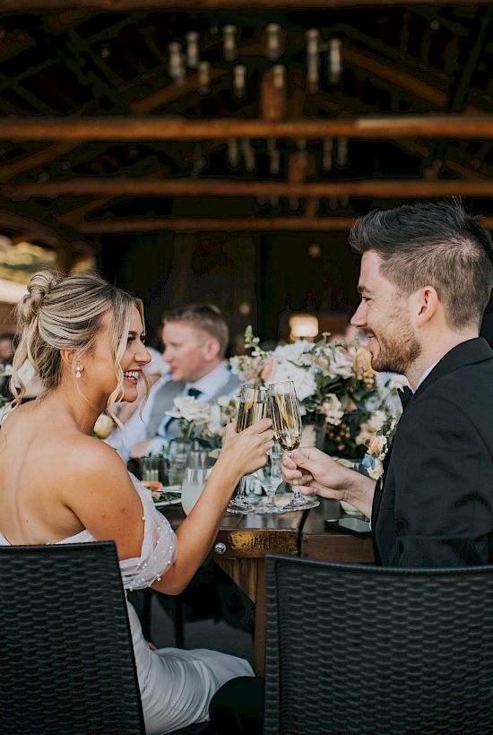 A wedding reception with a couple toasting amid a group of guests seated at a table outdoors.