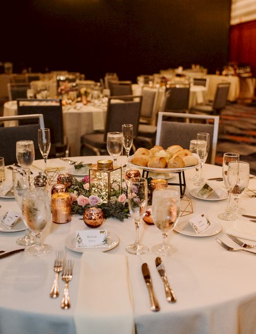 A wedding reception setup with elegantly arranged tables, white tablecloths, floral centerpieces, candles, and neatly placed tableware.