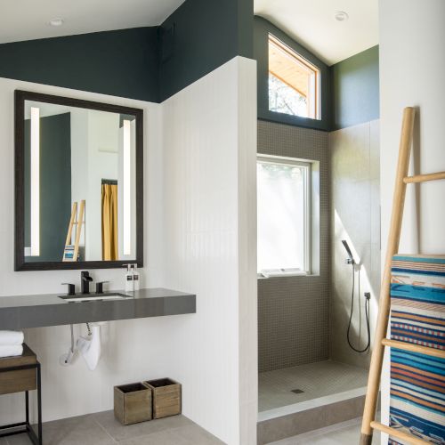 A modern bathroom with a shower, mirror, sink, ladder rack with towels, and natural light from a window.