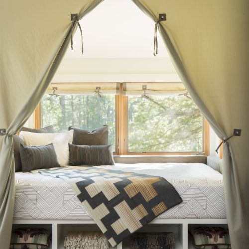 A cozy bed by a window with curtains, pillows, and a patterned blanket; storage shelves are underneath.
